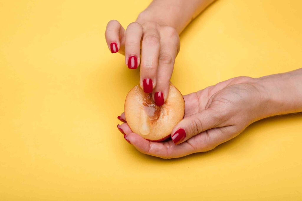 A photo with a yellow background showing a hand with red nails rubbing what would be the location of the clitoris on a peach. There is a liquid running down it.