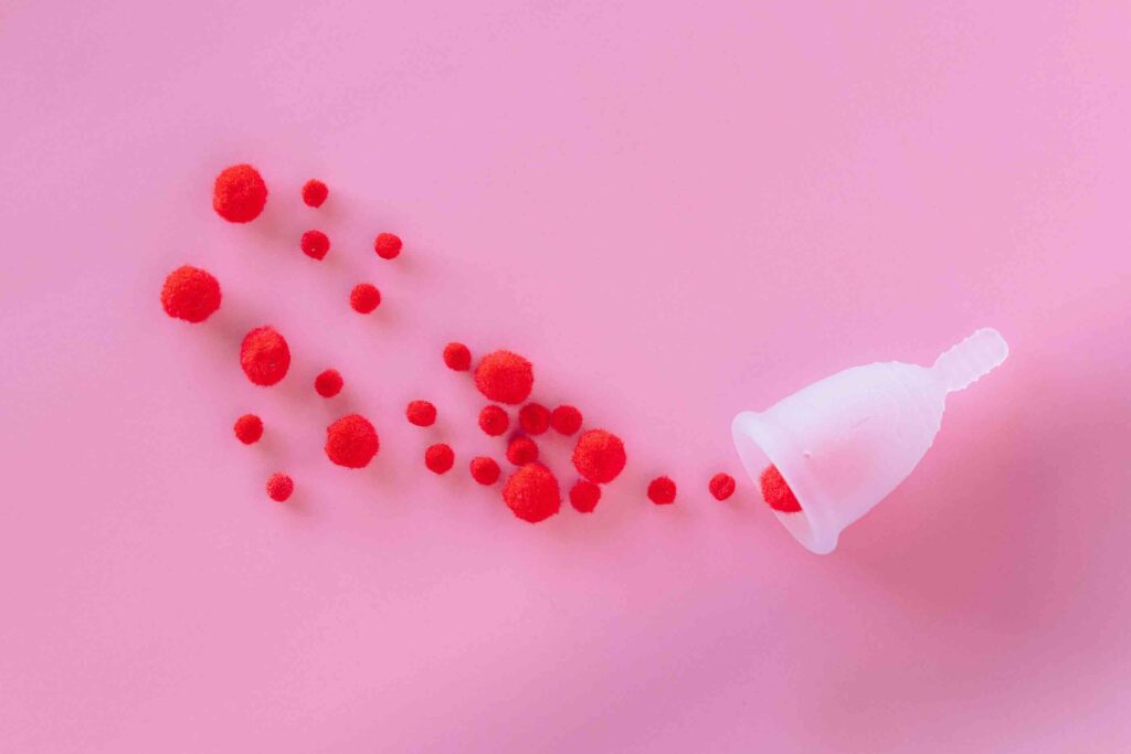 Image of a menstrual cup leaking red pom poms on a pink background