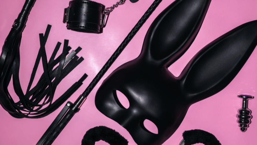 A photo of black adult toys including a leather bunny mask, whips and handcuffs on a pink background