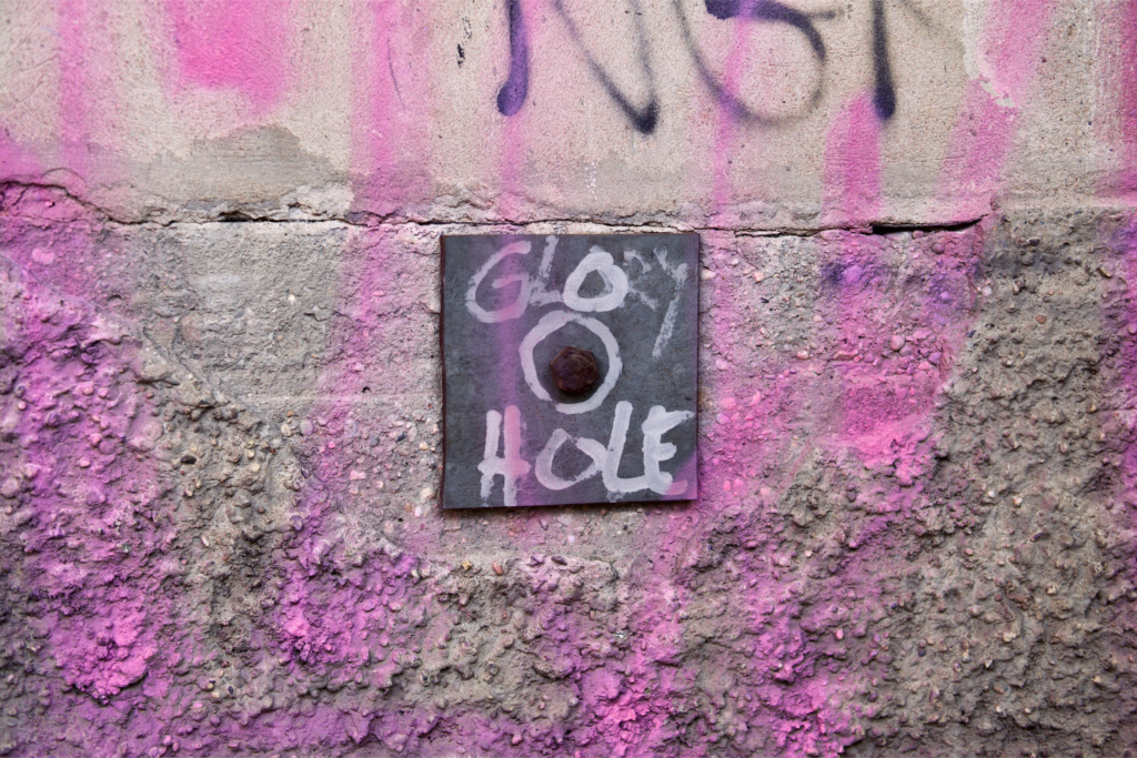 Photo of a hole in the wall with the writing glory hole around it with pink graffiti as it could be at a kinky party
