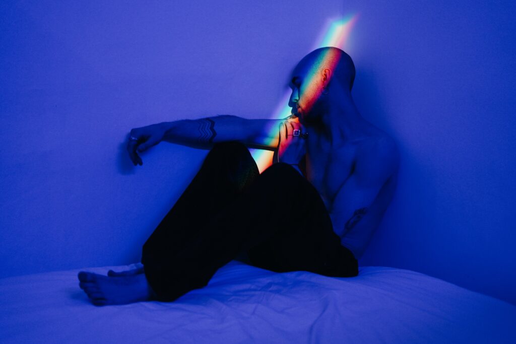 Photo of a man sitting with his head bowed in the corner with blue light and a rainbow light on his face. Picture by Cottonbro. It depicts a person struggling perhaps with their mental health.