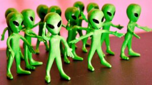 Small green toy alien figurines with large black oval eyes stand in a group, posed with their arms outstretched against a pink background.