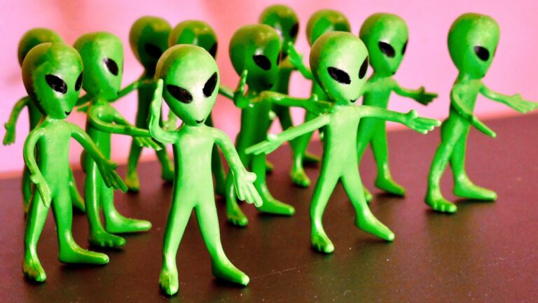 Small green toy alien figurines with large black oval eyes stand in a group, posed with their arms outstretched against a pink background.